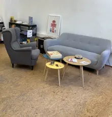 Sitzecke in der Werkstatt mit Sofa, Sessel und kleinen Tischen, dahinter Regale mit Bauteilen
